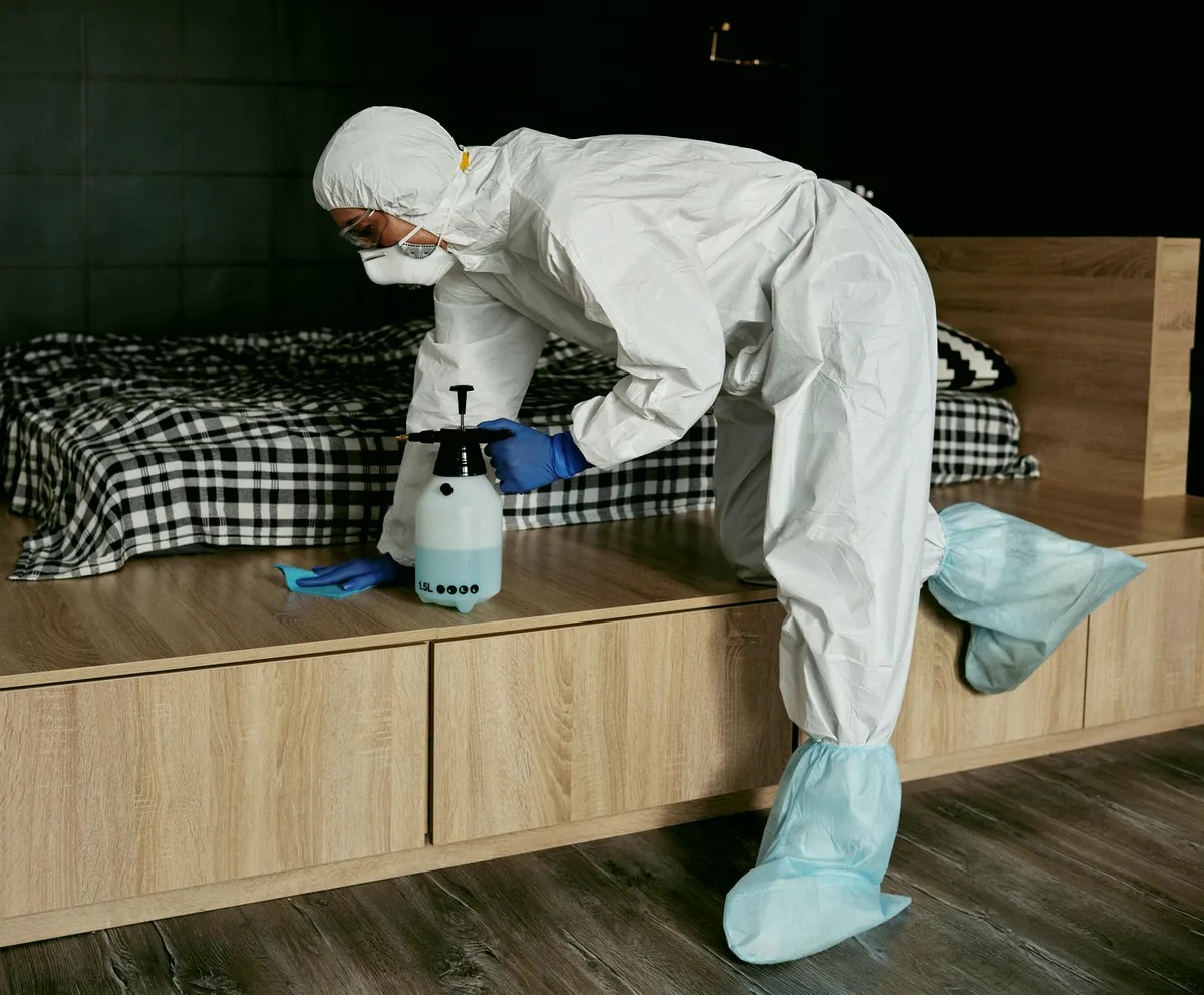 Técnica de dedetização com macacão, capuz e propés aplicando produto com pulverizador manual em volta da cama, no quarto